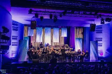 Koncert noworoczny na scenie MDK Lubliniec. Przed publicznością występuje Miejska Orkiestra Dęta. Scena oświetlona jest niebieskim kolorem.