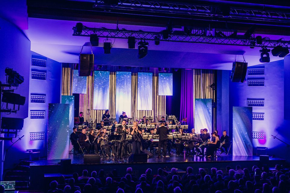 Koncert noworoczny na scenie MDK Lubliniec. Przed publicznością występuje Miejska Orkiestra Dęta. Scena oświetlona jest niebieskim kolorem.